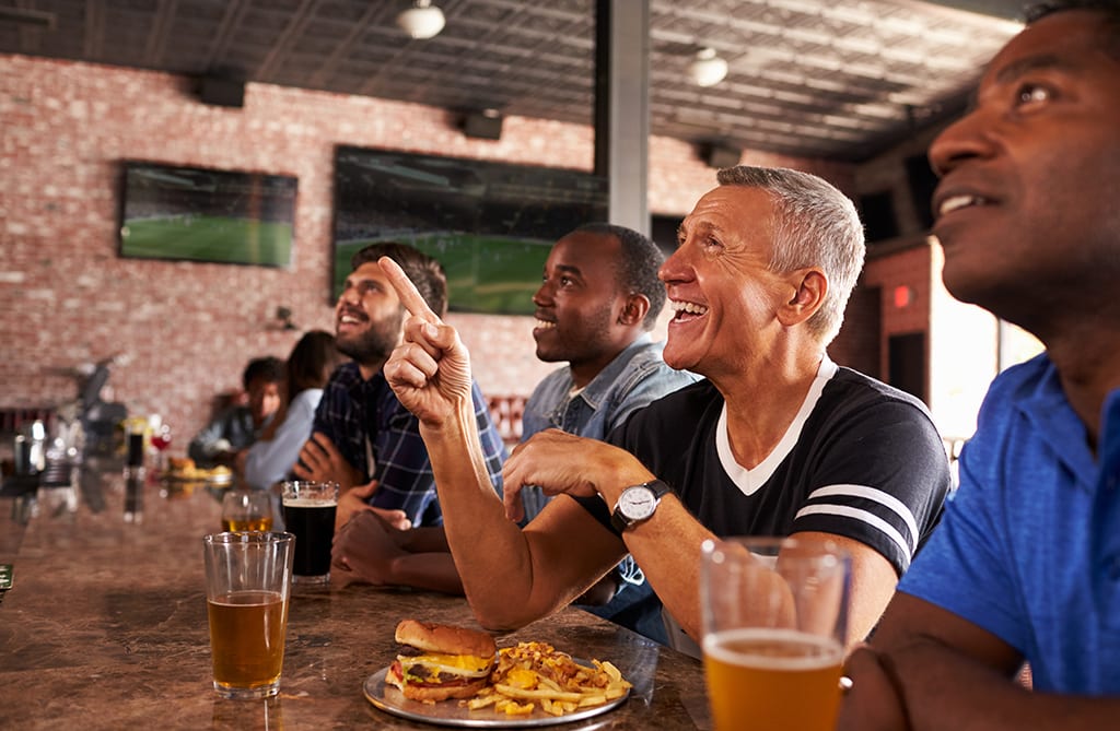 Male Friends At Counter In Sports Bar Watching Game