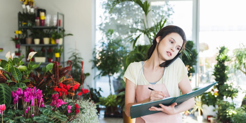 Young dedicated saleslady in floral shop taking orders over the phone in garden center and holding a notebook